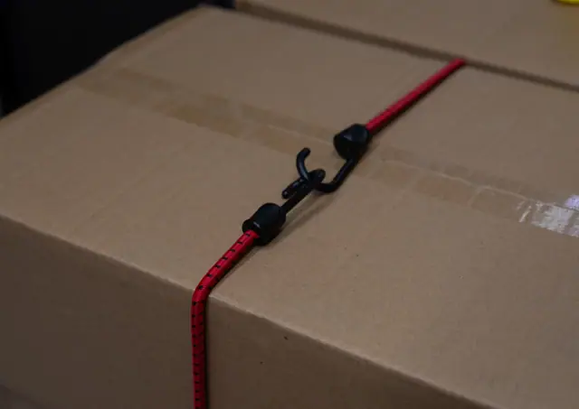 Red elastic strap running over the lid of a cardboard box, its hook in view
