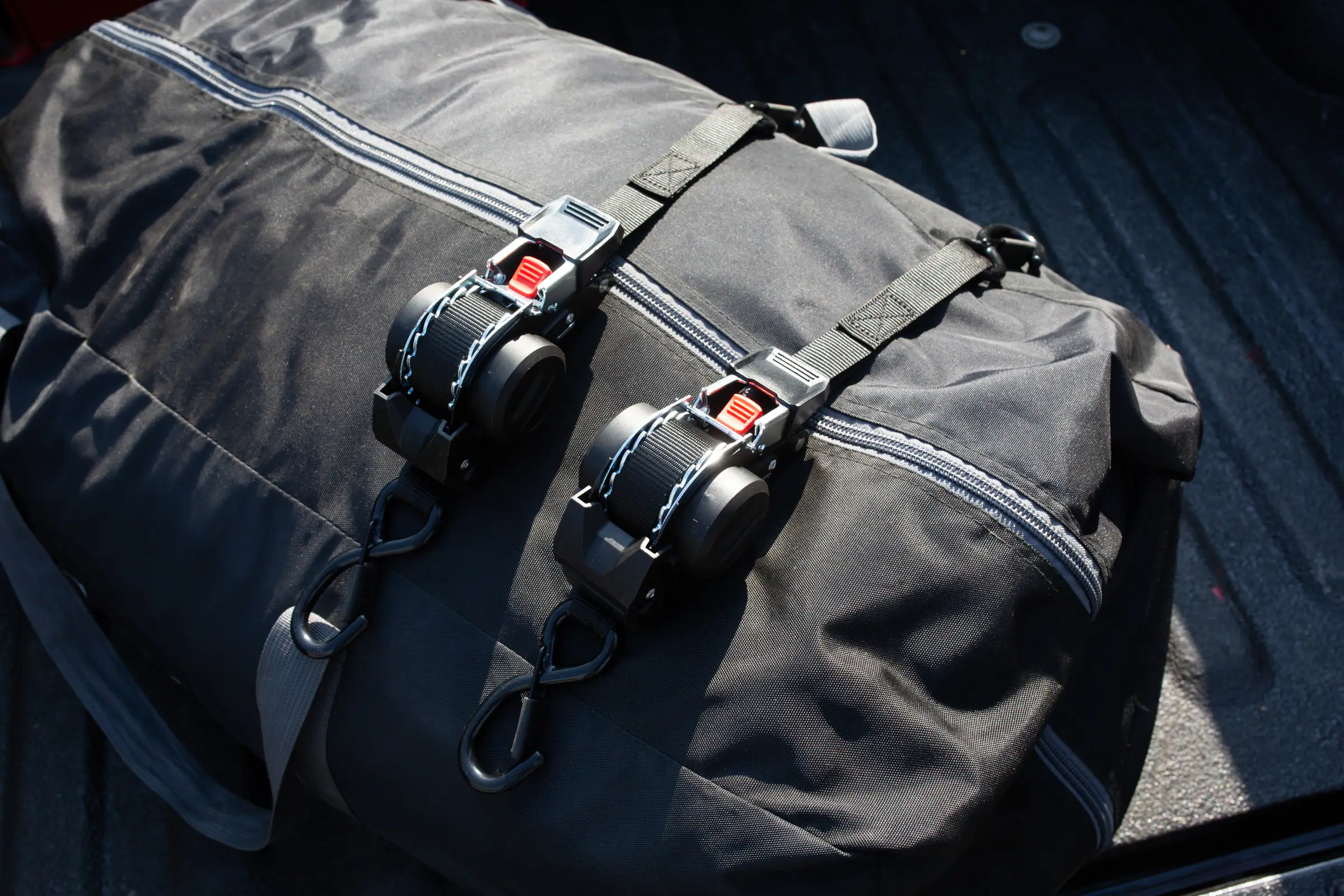 Two self-retracting ratchet straps cinched over a duffel bag in the bed of a pickup truck