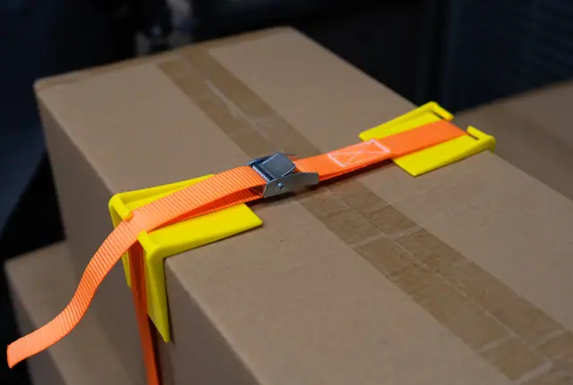 Cam buckle strap threaded through yellow ABS corner protectors on a carton