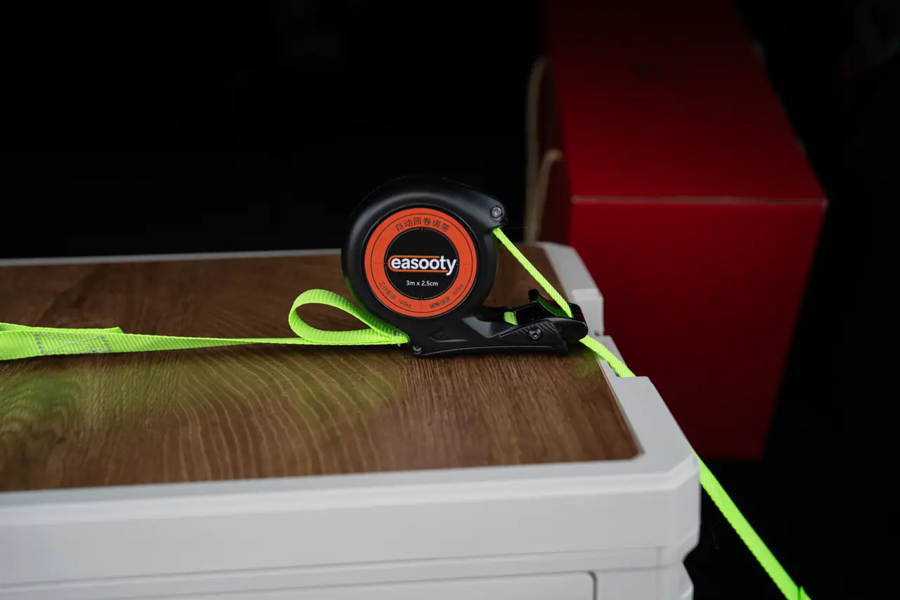 Auto-retract tie-down reel with its hook clipped to a crate edge and green webbing running across the load