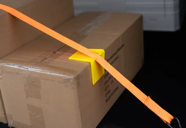A yellow ABS corner protector on a carton corner with an orange strap threaded through it
